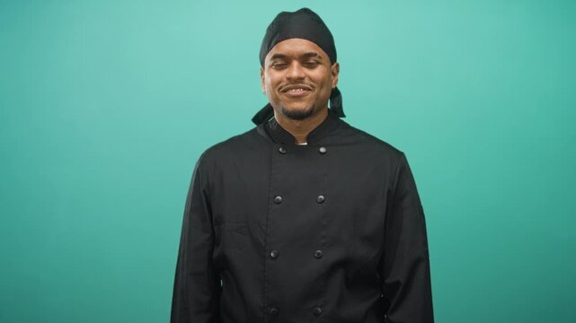 Man with beard smiling with eyes closed in chef uniform standing against teal studio backdrop; pride.