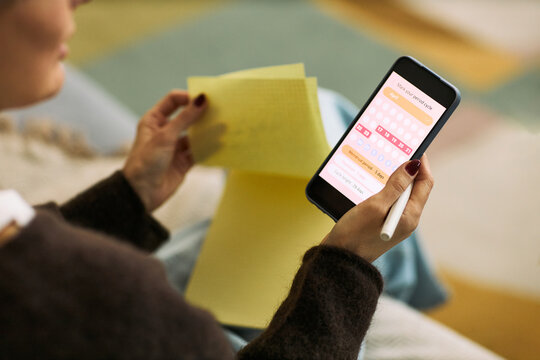 Caucasian woman holding smartphone displaying menstrual cycle tracking app while reviewing handwritten notes, using digital femtech solution for personal health management