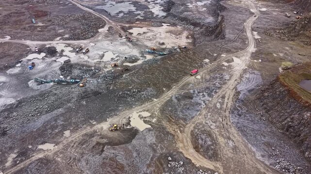 Heavy machinery works in a granite quarry. Trucks and equipment move around the site. The ground is rough and has stones everywhere. Various tasks are taking place in daylight.