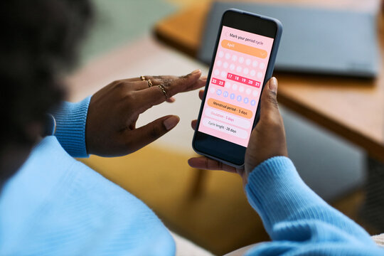 Black woman using smartphone tracking menstrual cycle with femtech health app, hand interacting with period calendar interface, focusing on reproductive health management
