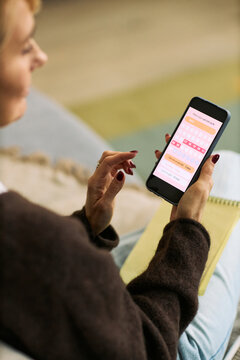 Middle aged Caucasian woman using smartphone health tracking app while sitting indoors, holding phone in one hand and notebook in other, focusing on femtech technology for wellness