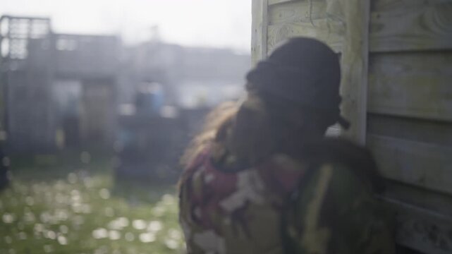 A person in camouflage and a mask takes aim with a rifle in a combat simulation game. Other players are positioned behind obstacles in an outdoor paintball arena.