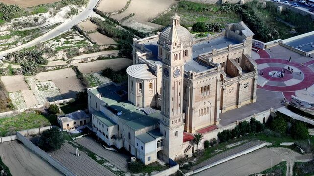 Aerial view of the majestic Ta Pinu Basilica in Gozo Malta during a bright sunny winter morning featuring golden limestone architecture against green terraced fields under a clear blue sky
