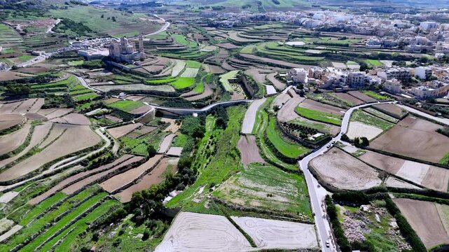 A wide high angle aerial drone shot captures the ancient Rotunda of Xewkija soaring above a lush green valley of terraced Mediterranean farmland in Gozo Malta during a bright sunny spring day