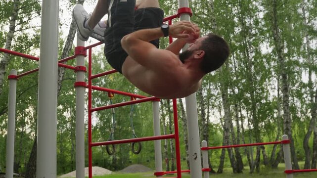 A strong, muscular man performs difficult inverted sit-ups while hanging upside down from a pull-up bar at an outdoor gym. Close-up footage of an intense calisthenics workout in a park.