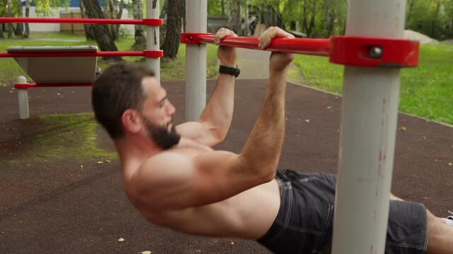 A shirtless, bearded, athletic man performs inverted rows (Australian pull-ups) on a low bar at an outdoor gym. The clip shows his muscular back and arms as he completes the bodyweight exercise.