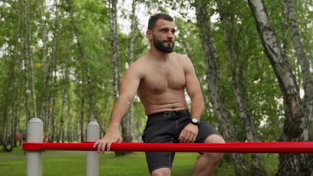 Shirtless bearded athlete performs dips on red parallel bars. Muscular man engaged in intense calisthenics workout in a green park. Footage of strength training and fitness lifestyle.