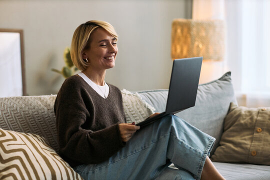 Caucasian mature woman sitting on sofa using laptop smiling during video call engaging with femtech health platform in home setting remote communication concept