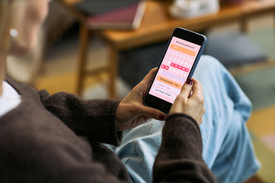 Caucasian woman holding smartphone using femtech health app for period tracking, sitting indoors focusing on menstrual cycle calendar interface on screen