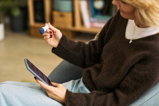 Middle aged Caucasian woman sitting holding smartphone in one hand and examining small medical device in other hand, engaging with femtech health technology at home