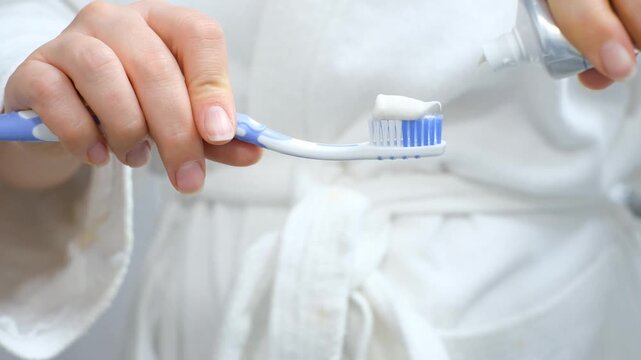 A woman applies toothpaste to a toothbrush against the background of bathrobe.