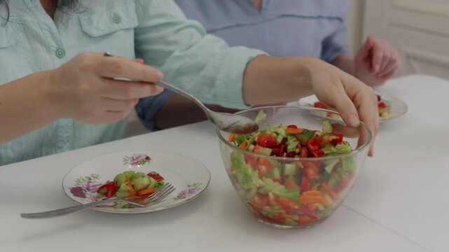 Close up of a woman's hands serving a fresh, colorful vegetable salad from a glass bowl onto a plate. Another woman eats in the background. Healthy eating and lifestyle concept.