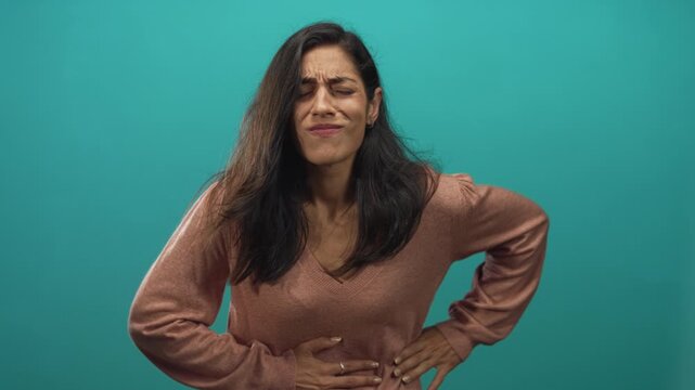 Young hispanic woman clutching stomach in teal studio, grimacing and pressing abdomen; discomfort nausea.