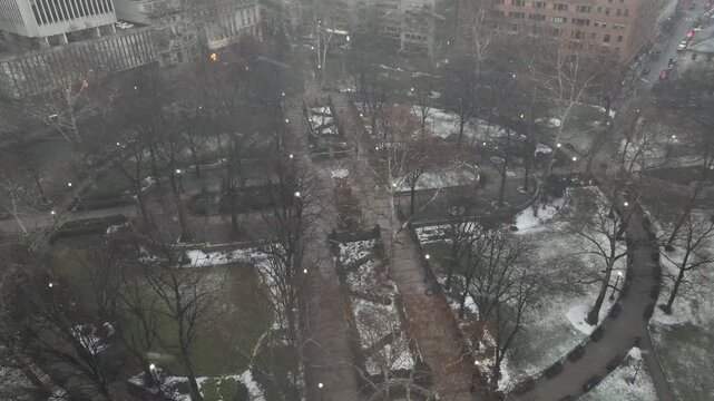 Rittenhouse Square in Philadelphia during gentle snowfall. Snow-dusted pathways curve through historic urban park, surrounded by city buildings and winter trees in Center City. Aerial Top down shot.