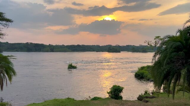 Golden sunset light reflects on the River Nile in Murchison Falls National Park, Uganda, with tropical riverbank vegetation and calm flowing water under dramatic clouds, static shot.