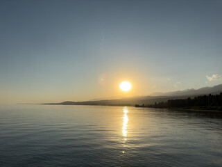 Obraz premium Scenic sunset over Issyk Kul Lake in Kyrgyzstan. The sun sets behind the mountains, casting a golden reflection across the water.