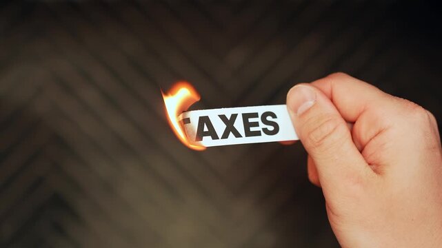 Close-up of a hand holding a paper label reading &ldquo;TAXES&rdquo; as the edge burns in a flame, symbolizing rising costs, tax burden, and financial stress.