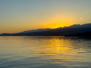 Obraz premium Scenic sunset over Issyk Kul Lake in Kyrgyzstan. The sun sets behind the mountains, casting a golden reflection across the water.
