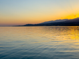 Obraz premium Scenic sunset over Issyk Kul Lake in Kyrgyzstan. The sun sets behind the mountains, casting a golden reflection across the water.
