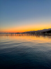 Fototapeta premium Evening sunset at Issyk Kul Lake in Kyrgyzstan. Long illuminated pier stretching into calm water with mountains on horizon.