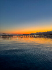 Fototapeta premium Evening sunset at Issyk Kul Lake in Kyrgyzstan. Long illuminated pier stretching into calm water with mountains on horizon.