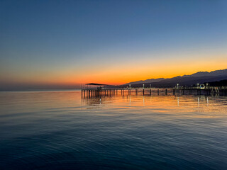 Obraz premium Evening sunset at Issyk Kul Lake in Kyrgyzstan. Long illuminated pier stretching into calm water with mountains on horizon.