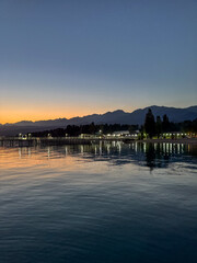 Fototapeta premium Evening sunset at Issyk Kul Lake in Kyrgyzstan. Long illuminated pier stretching into calm water with mountains on horizon.