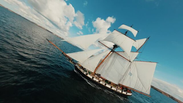 FPV drone flying around two-masted sailing ship cruising on blue ocean under sunny sky