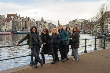 Mature women friends smiling and posing by an amsterdam canal, enjoying a winter city break together&mdash;carefree sightseeing, waterfront strolls, friendship and travel joy