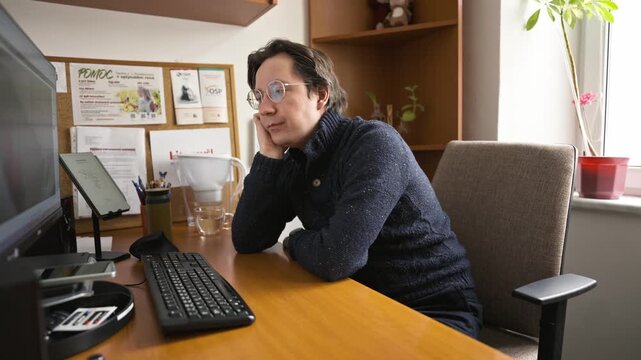 A man looking bored at his watch while sitting at a desk in front of a computer. Long working hours leading to fatigue and boredom in the office.