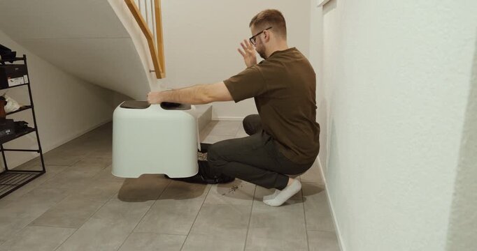 Young man kneeling next to a cat litter box in an apartment, reacting with disgust to bad odor. Concept of pet hygiene, unpleasant smell, cleaning routine, and domestic lifestyle challenges.