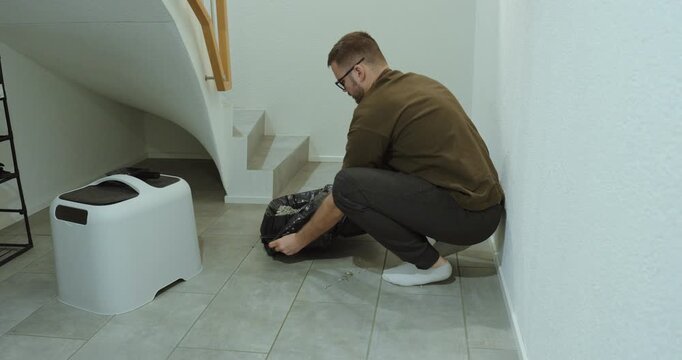 Young man kneeling under staircase while sealing and removing a trash bag from a cat litter box. Indoor pet hygiene routine showing responsible pet ownership and household cleaning process.