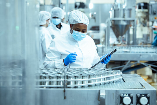 Female technician inspects stainless steel cans on conveyor belt in processing facility, writing notes for quality documentation and reinforcing food industry safety procedures.