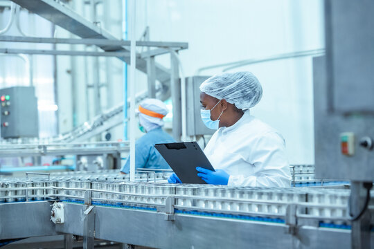 Industrial worker in hygienic clothing reviews canned products on conveyor system inside food manufacturing facility, documenting inspection results to ensure production compliance.
