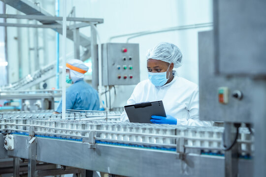 Female factory worker wearing protective uniform and face mask inspects canned food production line while checking data on clipboard in seafood processing plant, quality control and food safety.