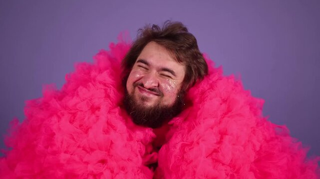 Horizontal 4K video. Young adult man with beard, glitter sparkles on male face, pink tulle fur coat, purple t-shirt looks at camera, smiles, dances, waves his arms, hand pistols on purple background