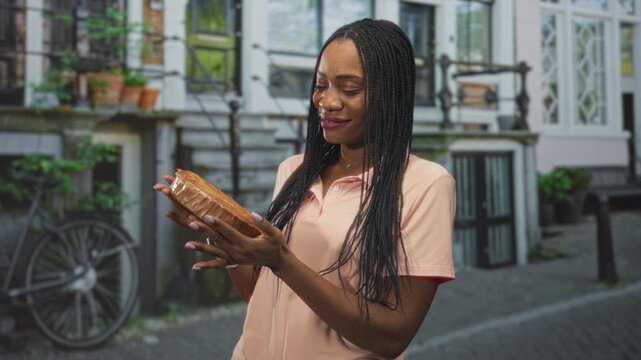 Woman holding packaged pastry in both hands, smiling as she inspects a round cake on a cobblestone street with bicycle and window pots; homemade baking contentment.