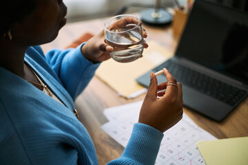 Young adult Black woman holding pill and glass of water near calendar and laptop, preparing to take medication, illustrating femtech health management and daily wellness tracking