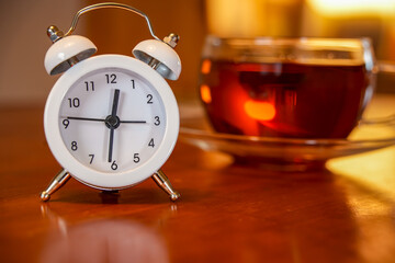 A white retro double bell alarm clock on a wooden table. In the background is a glass cup of black...