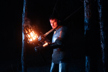 Obraz premium A knight in full armor, sword and torch in hand, walks through a gloomy night forest. A medieval warrior, bloodied and scarred, hunts and searches in the darkness.