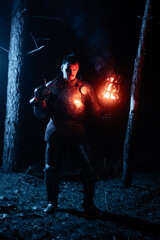 Obraz premium A knight in full armor, sword and torch in hand, walks through a gloomy night forest. A medieval warrior, bloodied and scarred, hunts and searches in the darkness.