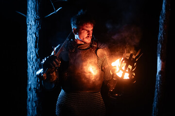Obraz premium A knight in full armor, sword and torch in hand, walks through a gloomy night forest. A medieval warrior, bloodied and scarred, hunts and searches in the darkness.