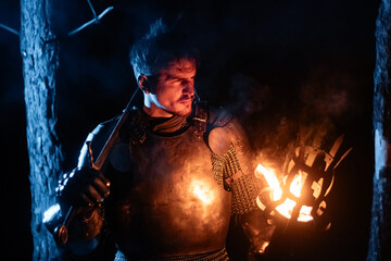 Obraz premium A knight in full armor, sword and torch in hand, walks through a gloomy night forest. A medieval warrior, bloodied and scarred, hunts and searches in the darkness.