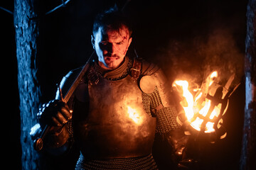 Obraz premium A knight in full armor, sword and torch in hand, walks through a gloomy night forest. A medieval warrior, bloodied and scarred, hunts and searches in the darkness.