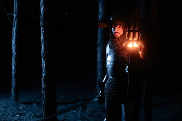 Obraz premium A knight in full armor, sword and torch in hand, walks through a gloomy night forest. A medieval warrior, bloodied and scarred, hunts and searches in the darkness.