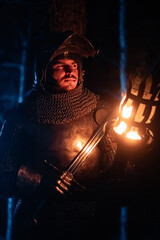 Obraz premium A knight in full armor, sword and torch in hand, walks through a gloomy night forest. A medieval warrior, bloodied and scarred, hunts and searches in the darkness.