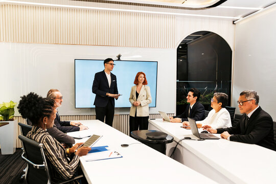 Professionals in a modern conference room deliver a presentation to colleagues, with laptops and tablets around a long table and a large display