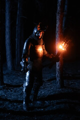 Obraz premium A knight in full armor, sword and torch in hand, walks through a gloomy night forest. A medieval warrior, bloodied and scarred, hunts and searches in the darkness.