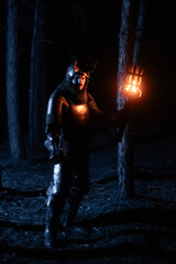 Obraz premium A knight in full armor, sword and torch in hand, walks through a gloomy night forest. A medieval warrior, bloodied and scarred, hunts and searches in the darkness.