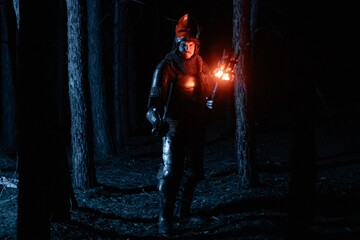 Obraz premium A knight in full armor, sword and torch in hand, walks through a gloomy night forest. A medieval warrior, bloodied and scarred, hunts and searches in the darkness.